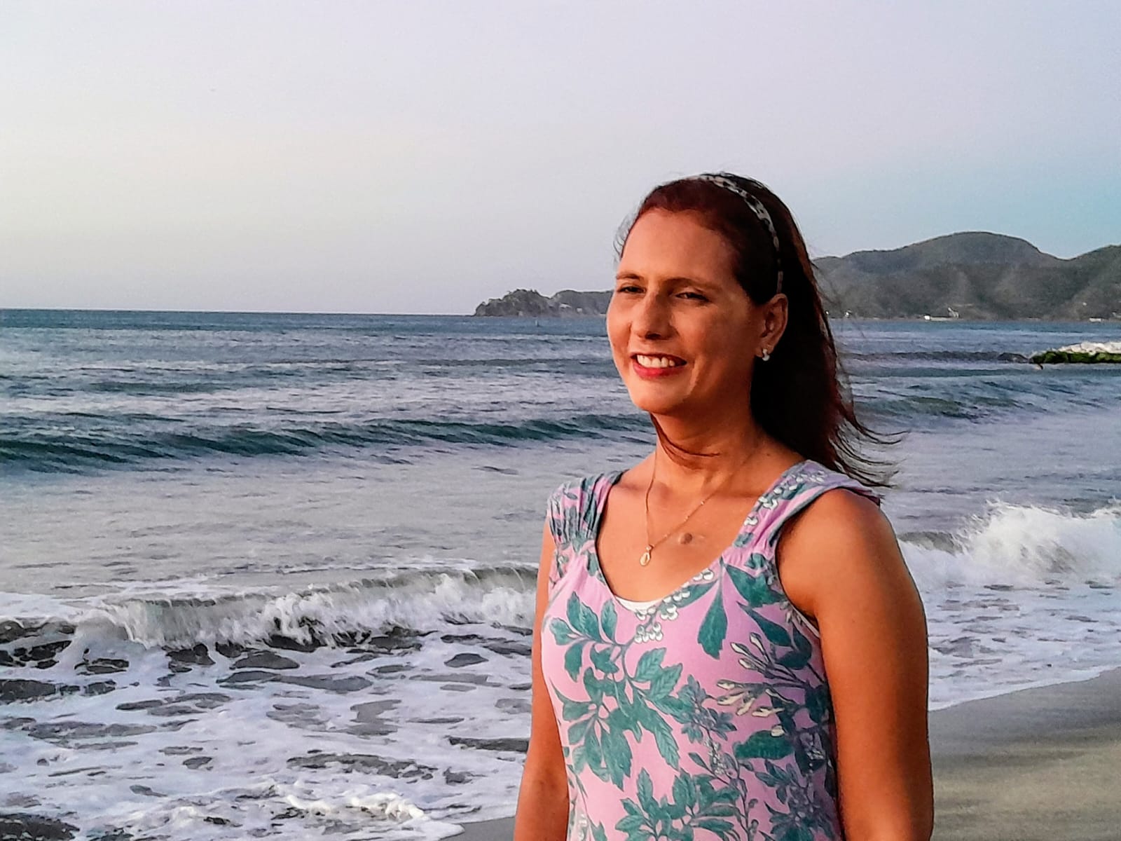 Sandra Cetina, vestida con un vestido de flores y con el pelo suelto, caminando a la orilla del mar mientras sonríe al horizonte 