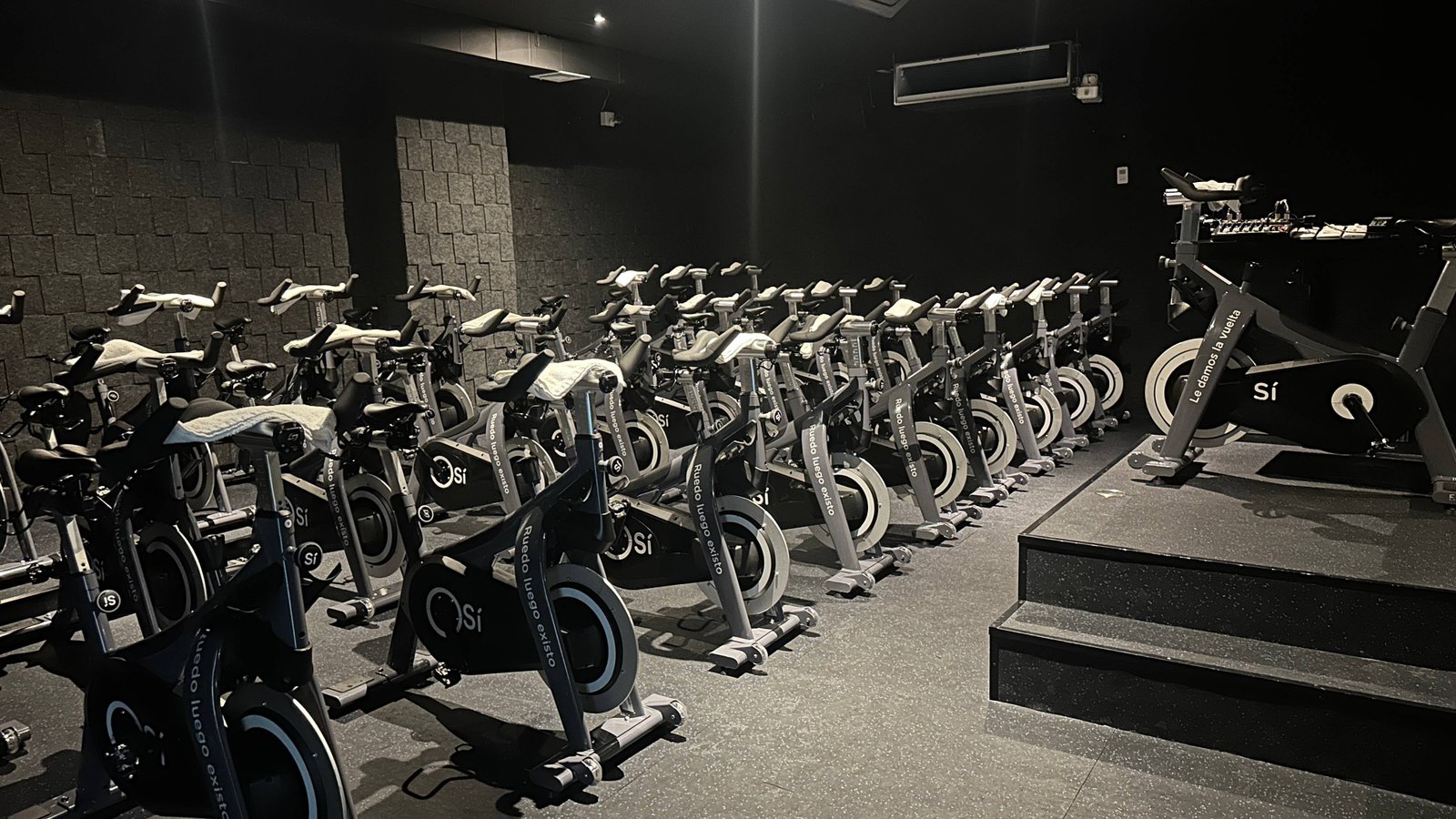 Salon de indoor Cycling. Se observan varias bicicletas estáticas organizadas en filas, todas del mismo modelo y color. Cada bicicleta tiene una toalla encima y manubrios con agarre ergonómico. 