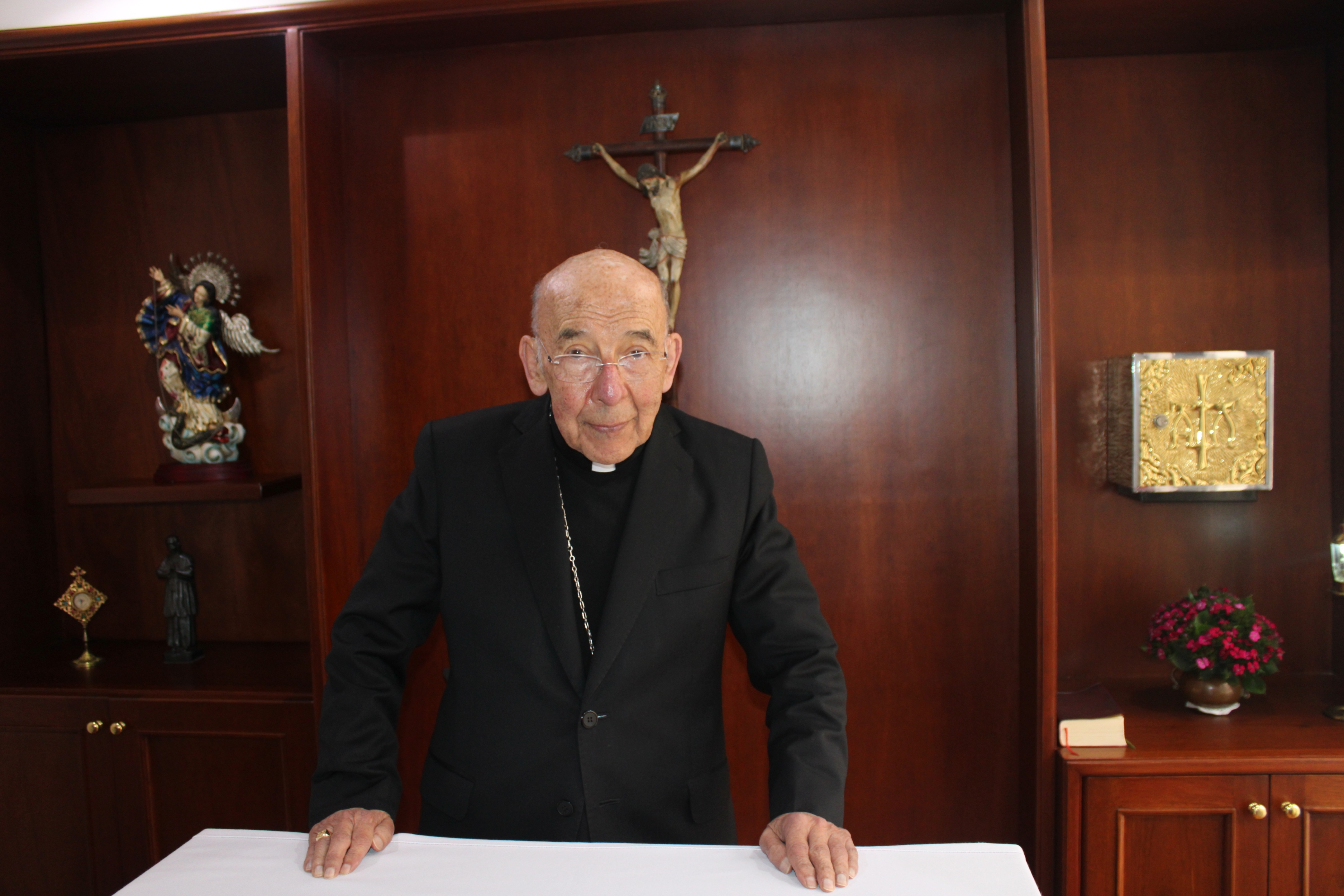 Monseñor Romero vestido con traje clerical negro, de pie frente a una mesa, con un crucifijo en la pared y figuras religiosas a su alrededor.