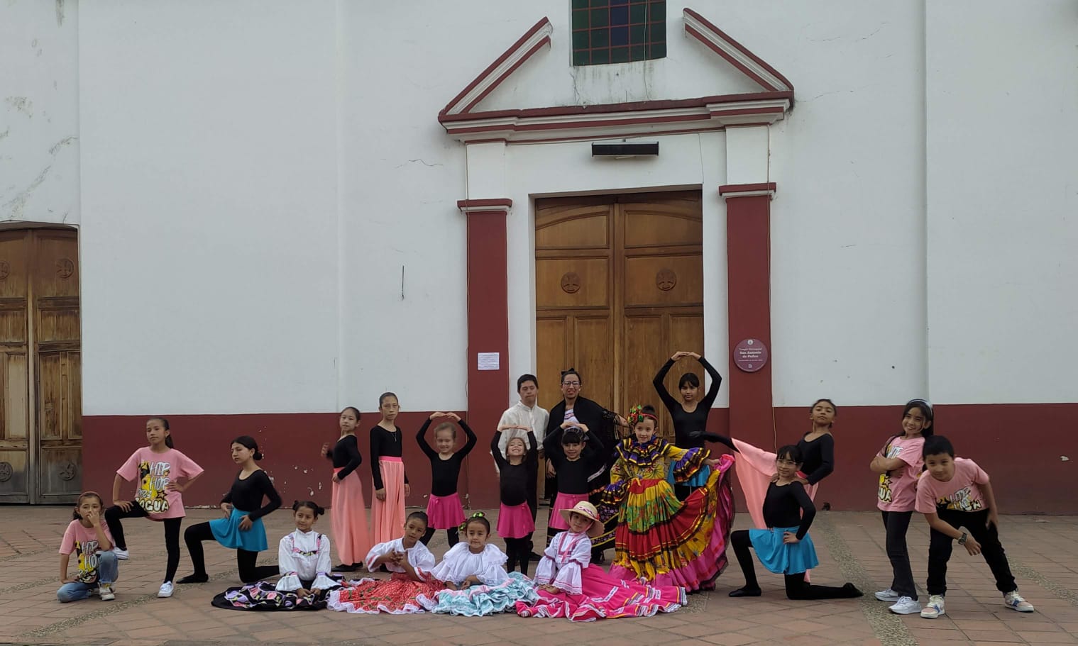 Grupo de niños y niñas de la Casa de la Cultura de Cogua posan frente a la iglesia del municipio, vestidos con trajes típicos coloridos y atuendos de danza contemporánea, representando la diversidad artística de la región.