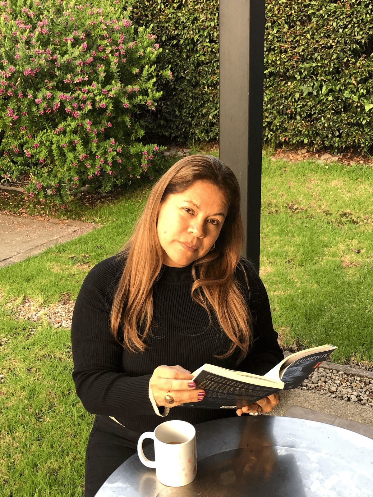 Leslly Romero aparece sentada en un espacio al aire libre, rodeada de vegetación. Con un libro entre las manos y una taza de café sobre la mesa, transmite una escena de serenidad y concentración. La luz natural ilumina el entorno, reforzando la idea de un momento íntimo de lectura y reflexión en medio del jardín.