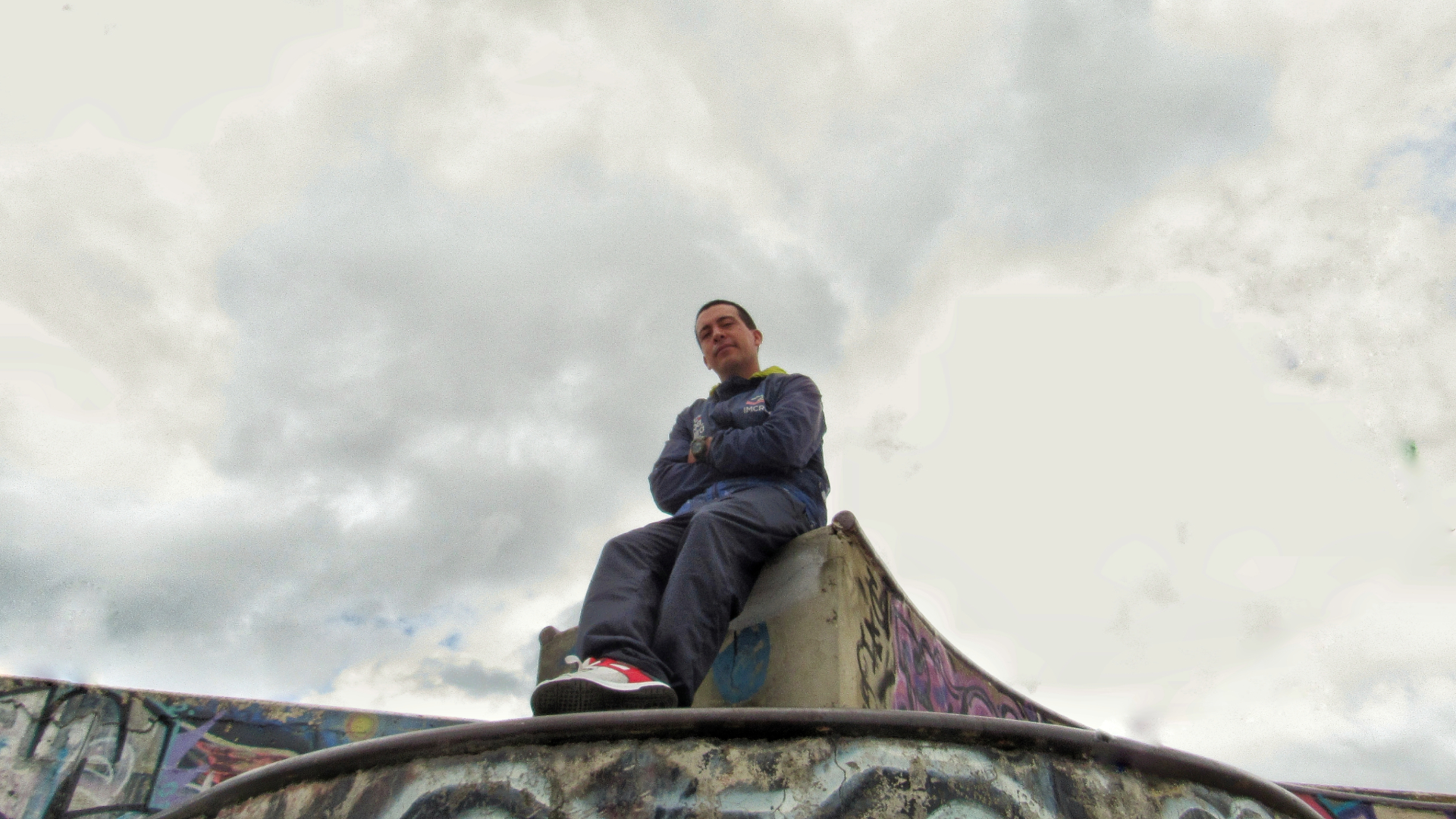 En plano contrapicado, se encuentra en el centro de la imagen al profesor Julian Pedraza. Este porta su uniforme de instructor y se encuentra posando en la barandilla de un skatepark.