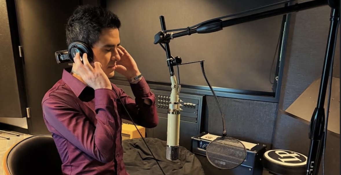 Diego Caceres se encuentra en su estudio de grabación profesional, sentado frente a un micrófono de condensador mientras usa audífonos para monitorear el sonido. Lleva una camisa color vino y botas marrones, concentrado en el proceso de grabación o mezcla musical. El ambiente moderno y bien equipado refleja un espacio creativo dedicado a la producción de audio. 