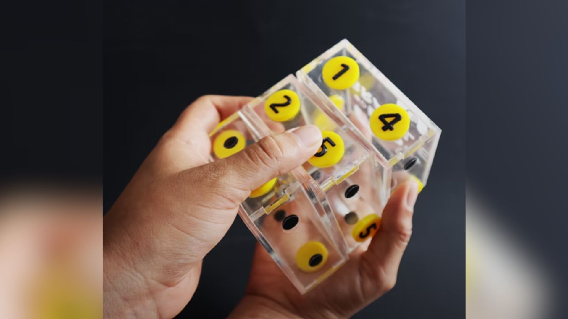 Elemento didádicto en forma de cubo de cristal con números de color verde, rodeados de un contorno amarillo, que es manipulado por dos manos humanas.