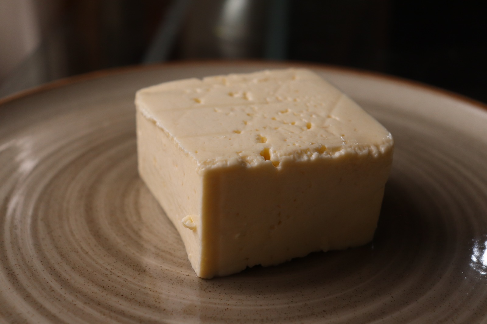 Bloque de queso blanco de forma rectangular sobre un plato de cerámica de color crema, iluminado con luz cálida y fondo desenfocado