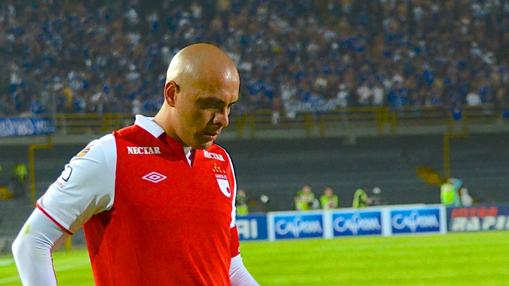 En la fotografía se ve a Omar Pérez en el año 2012. Se encuentran en el estadio El Campin, en la ciudad de Bogotá. Pérez es un hombre sin cabello y viste una camisa de color rojo en torso y blanco en las mangas, correspondiente al equipo de fútbol Santa Fe. Pérez se encuentra en la cancha del estadio. De fondo se ve a una aglomeración de espectadores, en la parte superior, sentados en las graderías del estadio. En la parte baja se identifican a policías y personal de seguridad.