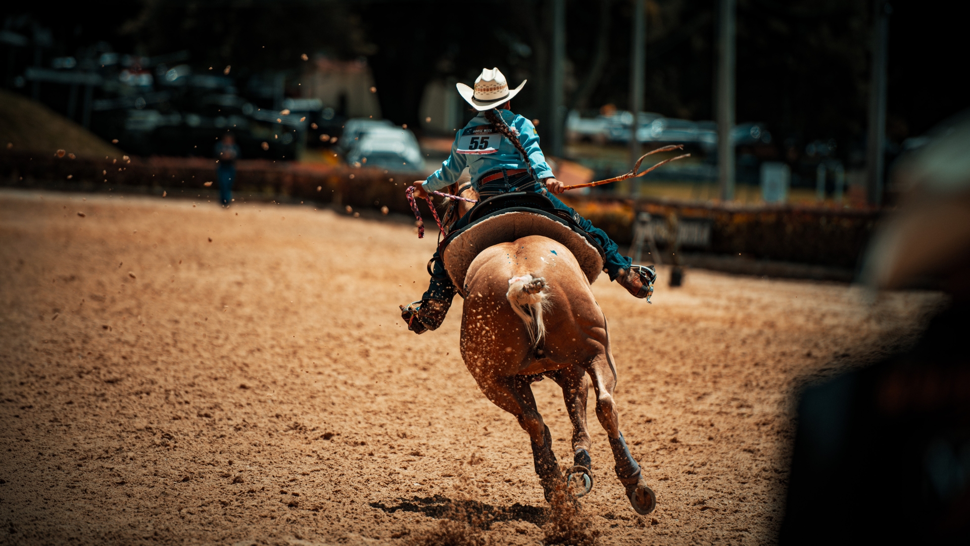 En una pista de tierra, una mujer con sombrero, camisa larga, pantalones y botas vaqueras, cabalga sobre un fuerte caballo, a gran velocidad e inclinados levemente. 