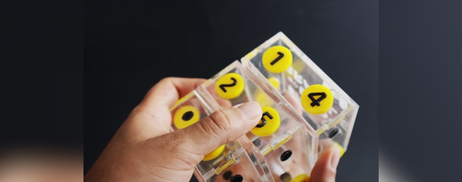 Elemento didádicto en forma de cubo de cristal con números de color verde, rodeados de un contorno amarillo, que es manipulado por dos manos humanas.